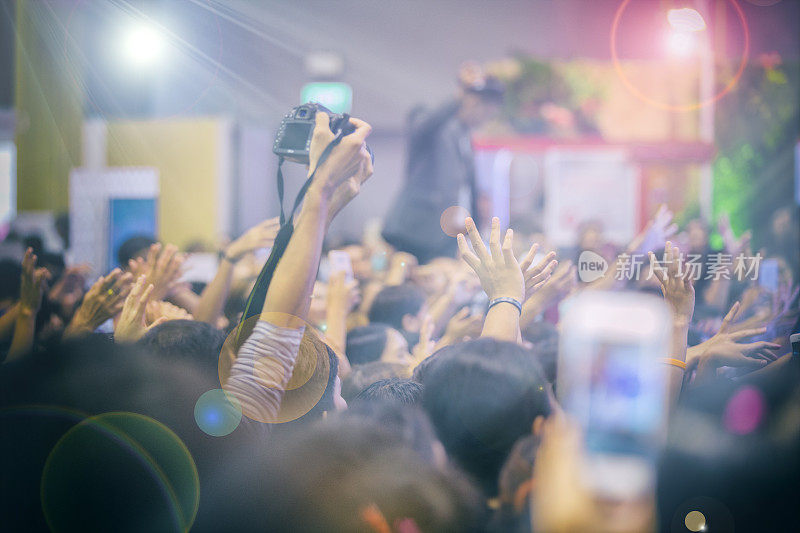 亚洲女性拿着相机在演唱会舞台灯光下人群中拍照，背景模糊