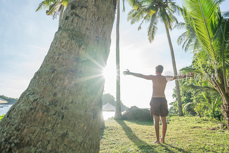 年轻男子在棕榈树之间的手臂在印度尼西亚巴厘岛在日落凝视他周围的自然-人们度假异国情调的地方概念