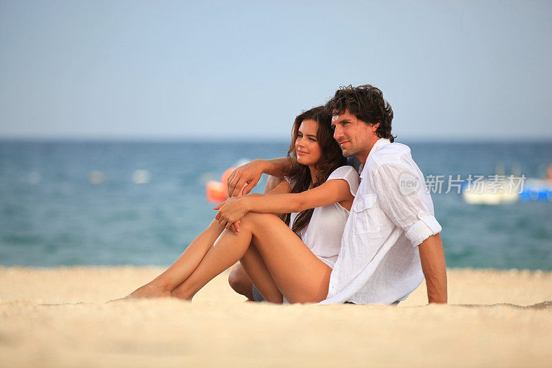 快乐的年轻夫妇坐在海滩和愉快和微笑，而日光浴