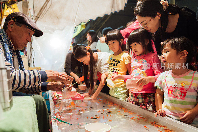 一位日本老人把金鱼装进塑料袋送给女孩，而其他女孩则在泡菜摊上玩传统的挖金鱼游戏。