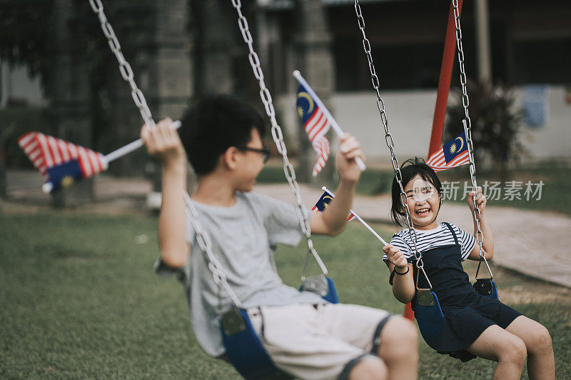 一对亚洲华裔兄妹手捧马来西亚国旗，在儿童操场上荡秋千，微笑着享受亲密时光