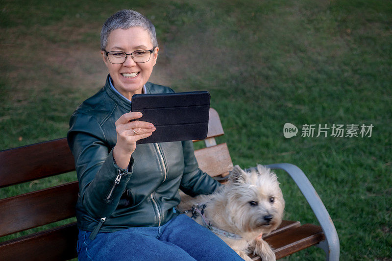 一个成熟的女人和一只狗坐在户外的长椅上，用平板电脑和朋友视频通话