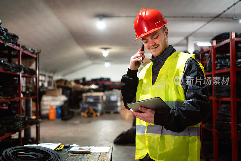 一名工厂工人的男性工程师在仓库里一边用智能手机与客户通话，一边使用数字平板电脑