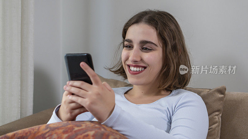 年轻女孩坐在沙发上微笑着看着手里的手机。主动给所爱的人发短信的女孩的幸福。
