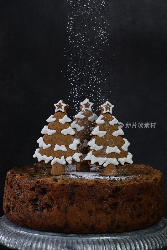 在自制的圣诞水果蛋糕上筛上糖霜的图像，上面是用白色皇家糖霜覆盖的姜饼圣诞树饼干，放在银色的蛋糕架上，黑色的背景，重点放在前景上