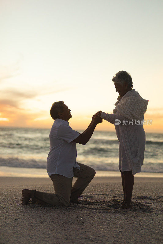 在海滩上跪着求婚的年长混血儿男人手牵着手的侧视图