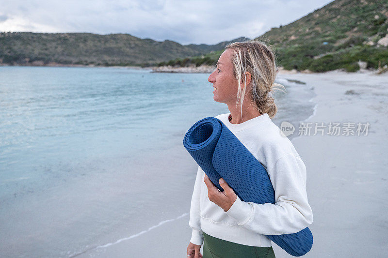 运动的年轻女子准备锻炼，她走在海滩上，在日出时，她的瑜伽垫看向大海