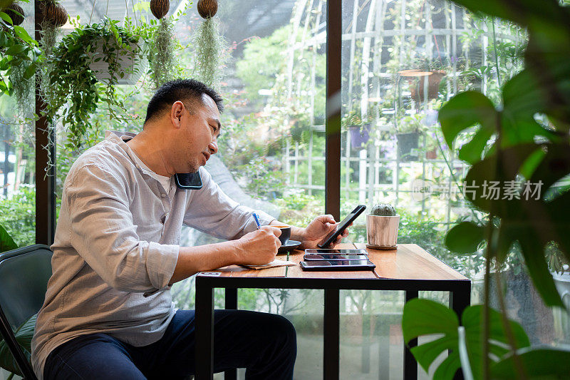 中年亚洲男子在咖啡馆打电话，一只手拿着笔，一只手拿着另一只手机
