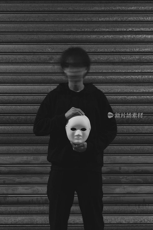 万圣节的肖像，一个年轻的黑衣人拿着一个白色的面具，他的脸模糊地站在一个金属门前