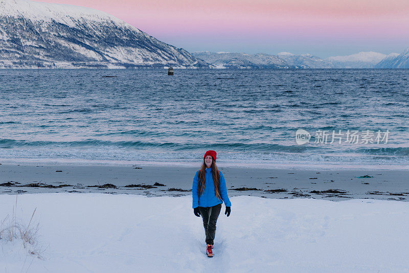 快乐的女人背着背包走在冬天的海滩上，在挪威的粉红色日落期间，白雪覆盖的山景
