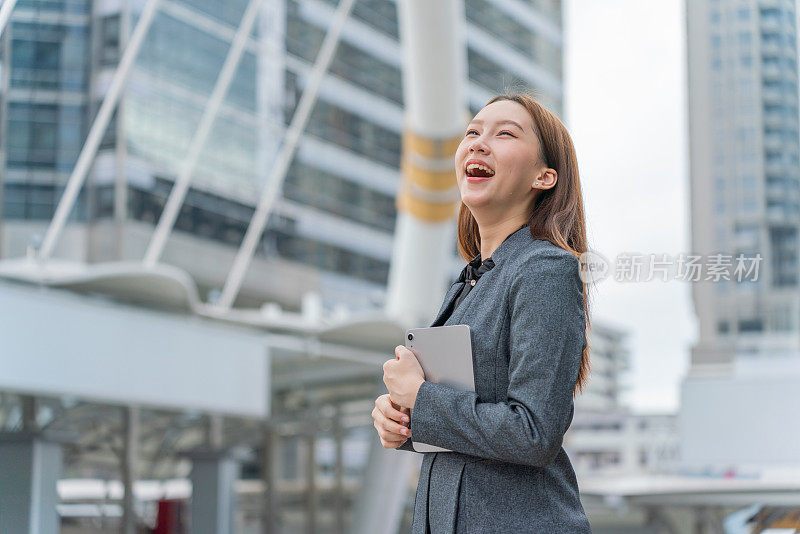 一名年轻的亚洲女商人穿着灰色的办公套装，在商业区使用她的平板电脑时微笑着