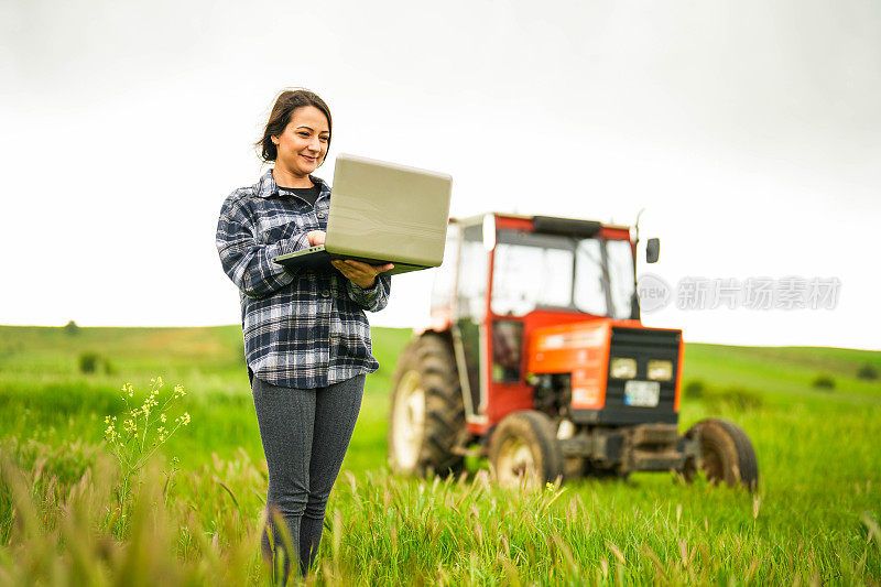 年轻的女农学家站在拖拉机前，用笔记本电脑控制麦田，智能耕作方法