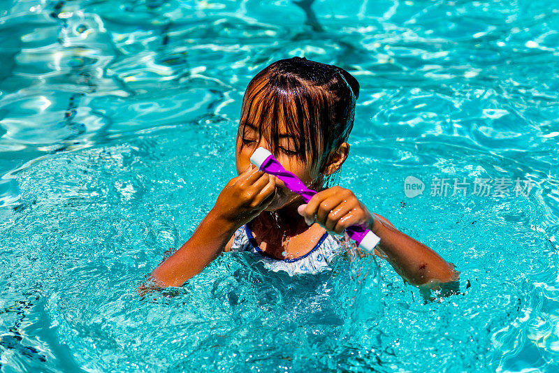 夏日里，一个大约7岁的小女孩沐浴在温暖的阳光下，在泳池里享受着纯粹的快乐。当她拿着潜水棒时，空气中充满了她戏谑的水花和笑声。