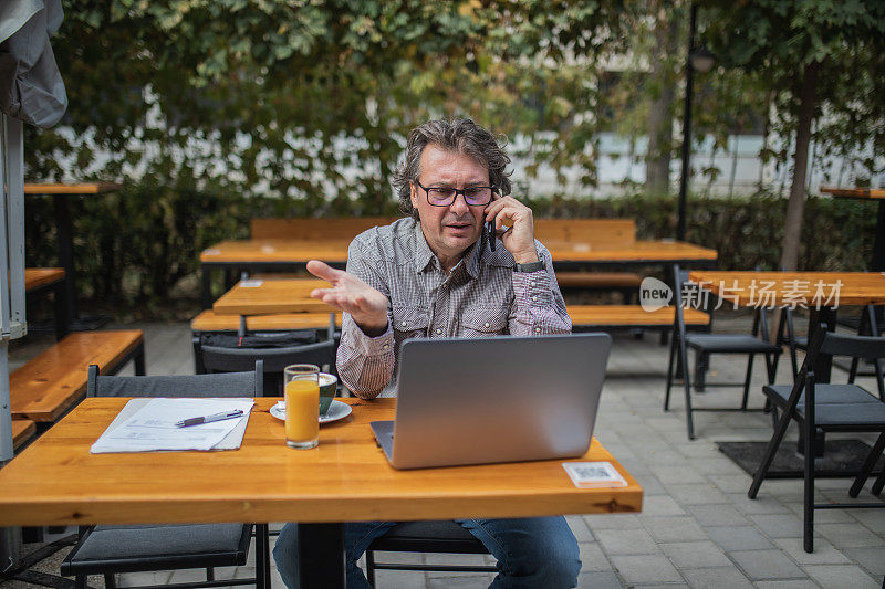 一个成熟的中年男子坐在咖啡厅里，一边用笔记本电脑工作，一边用手机聊天。