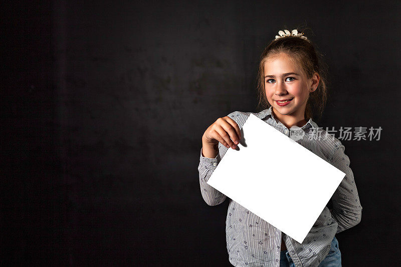 微笑的小女孩拿着假的白色海报空白框，在黑色背景下摆姿势，看着相机。少儿宣传空模板，空白海报在手。广告的概念。复制广告文本空间