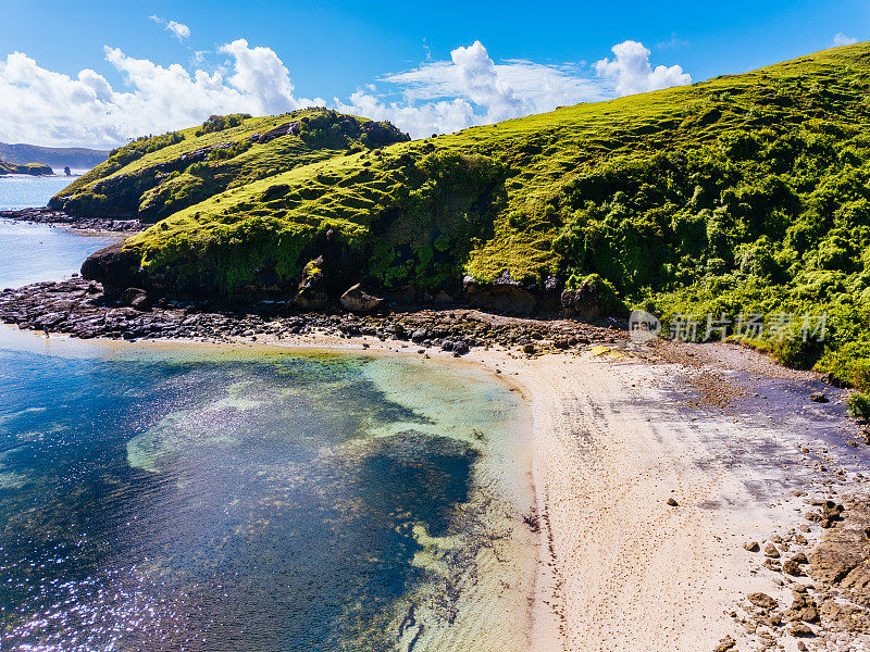 海岸景观:龙目岛，生机勃勃的绿色山坡，清澈的蓝色海水，明亮的天空和蓬松的云下的沙滩海岸线。