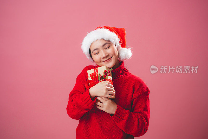 亚洲女人戴着圣诞老人的帽子，拿着礼盒开心地微笑，孤立在粉红色的背景上。