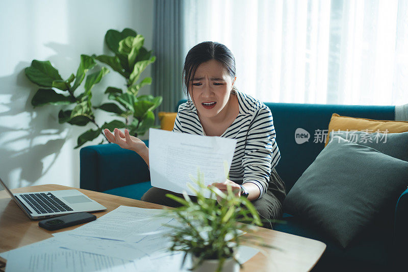 亚洲女性坐在沙发上，担心银行票据、抵押贷款债务和生活成本的账单支付问题。