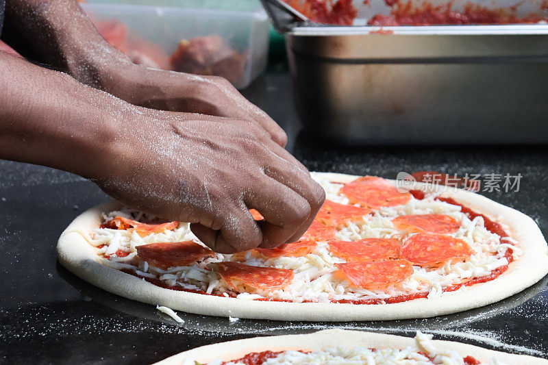 难以辨认的Pizzaiolo(披萨厨师)在未煮熟的披萨基础上添加意大利辣香肠片的特写图像，意大利辣香肠披萨富含番茄马里纳拉酱，磨碎的马苏里拉奶酪，商业厨房设置，重点放在前景