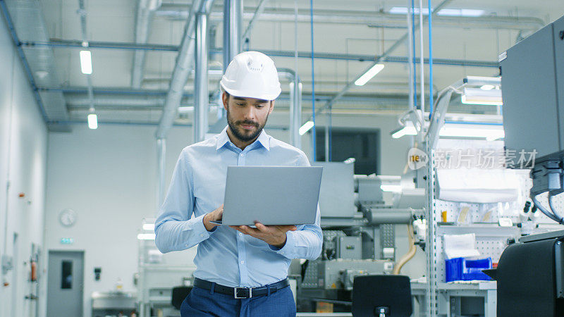 戴安全帽的总工程师手拿笔记本电脑穿过轻型现代工厂。现代工业环境下的成功英俊男人。