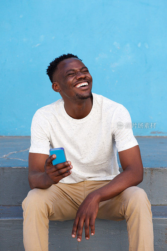 一个兴高采烈的非洲年轻人坐在台阶上，手里拿着手机