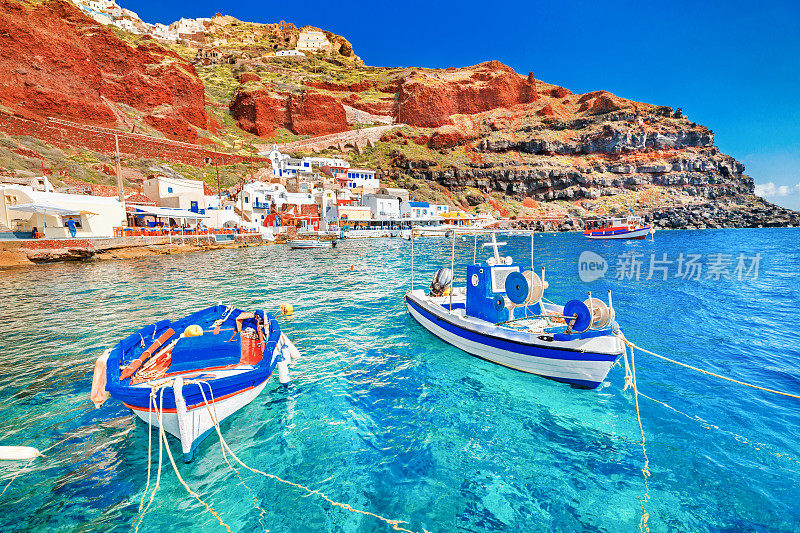 希腊。在爱琴海的希腊圣托里尼岛的伊亚伊亚村，两艘渔船停泊在迷人的蓝色水域的码头上，这是令人惊叹的古老港口全景。