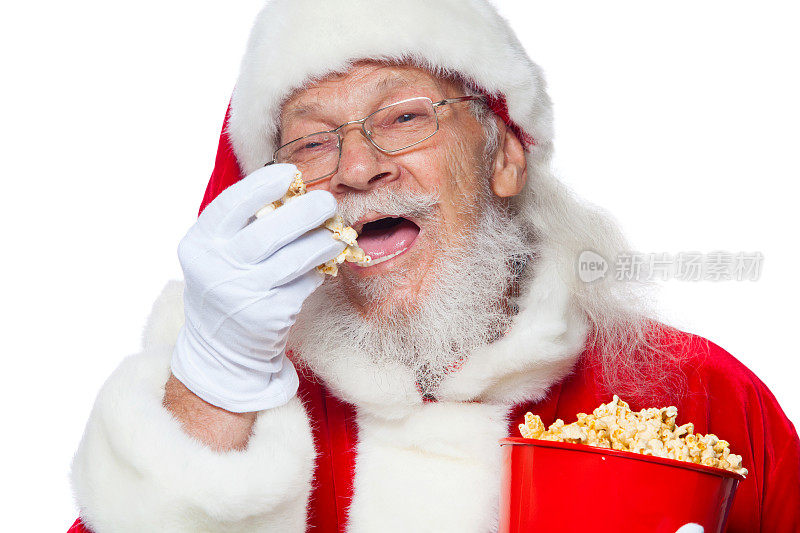 圣诞节。善良的圣诞老人面带微笑，戴着白手套，嘴巴张着，一只手拿着一个装着爆米花的红桶，拿了一些爆米花，准备用第二只手吃它。参观电影院的概念，用爆米花看电影。隔离