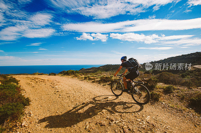 一个年轻人在西班牙的一条自行车道上骑着山地车，路上的背景是地中海。戴着头盔，背着黑色的背包