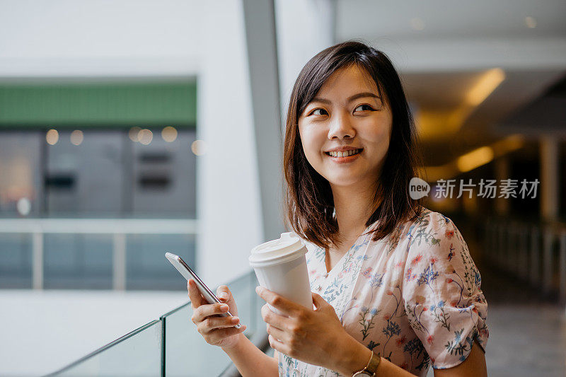 快乐的年轻亚洲女性使用智能手机和拿着纸咖啡杯