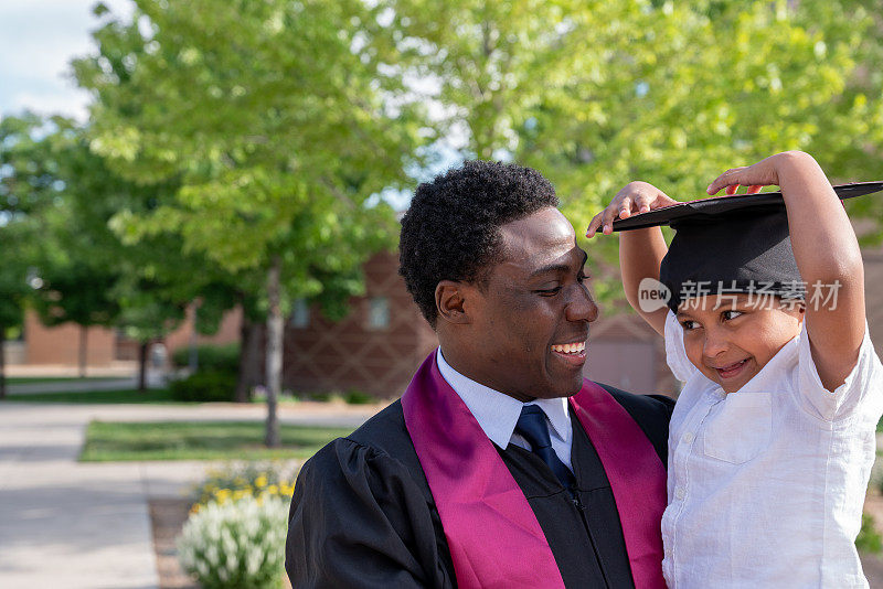 非裔美国男大学毕业生戴着帽子和长袍，在毕业典礼上和他三岁的儿子一起玩