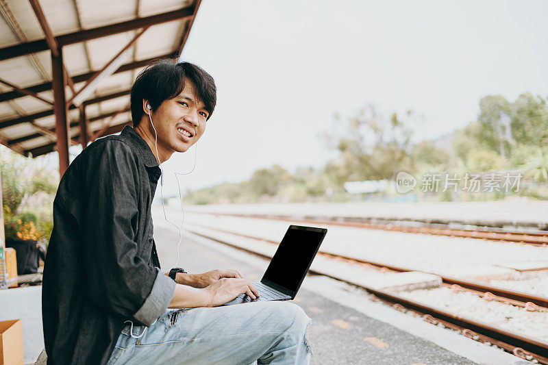 年轻的亚洲人使用笔记本电脑工作和搜索位置的旅行在火车站。自由职业者工作和旅行的概念。