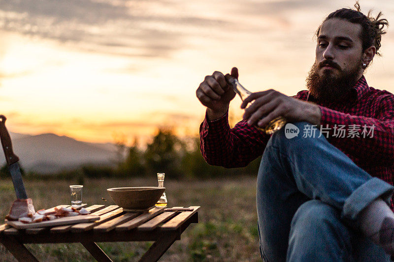 一个在山上的男人，一个伐木工人，喝着自制的白兰地，吃着自制的食物，而在他身后是美丽的日落，休息辛苦的工作。