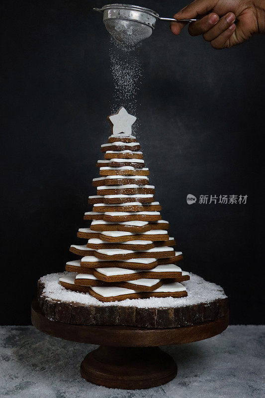 无法辨认的人在圣诞树上筛糖粉的图像，圣诞树是由堆叠的姜饼星饼干和白色皇家糖霜组成的，木制蛋糕架，人造雪，黑色背景，聚焦前景