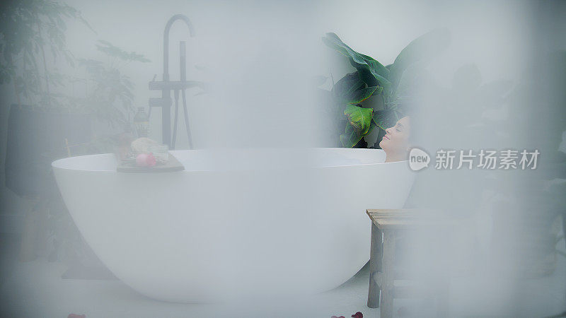 美丽的女人在现代浴室放松。在有热带植物的豪华浴室里听音乐