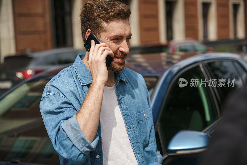 一位留着大胡子、穿着牛仔裤衬衫的出租车司机靠在一辆汽车上，面带微笑地讲着电话