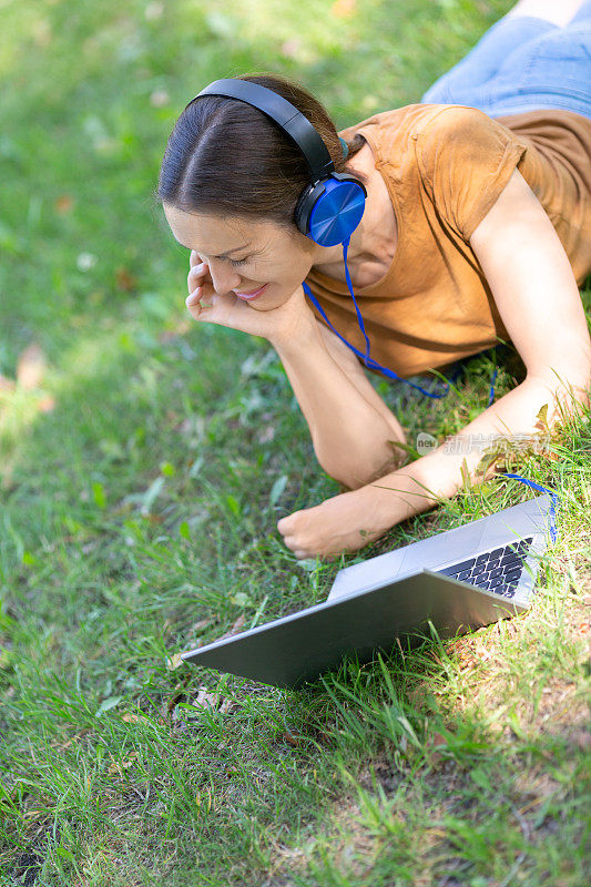 远程商务，远程学习。白种人美女戴着耳机躺在草地上，用笔记本电脑工作在公园的户外，夏天，垂直拍照，选定焦点。