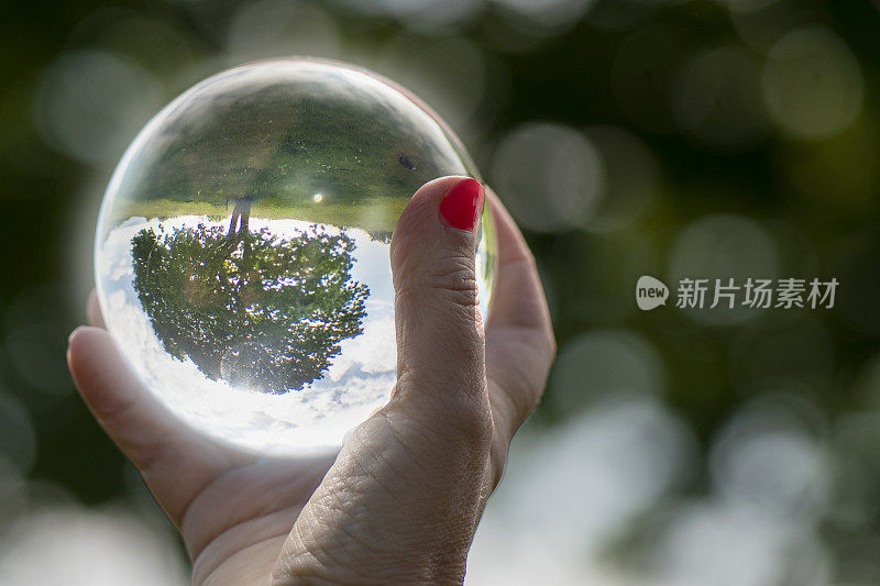 关注女性照顾自然和气候。一个女人的手拿着一个红色指甲的水晶球，自然映照在上面。