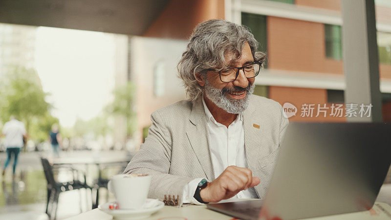 成熟的商人，留着胡子，戴着眼镜，穿着灰色夹克，坐在咖啡馆里喝咖啡。中年经理，成功的男人在笔记本电脑上工作，坐在户外的咖啡馆里