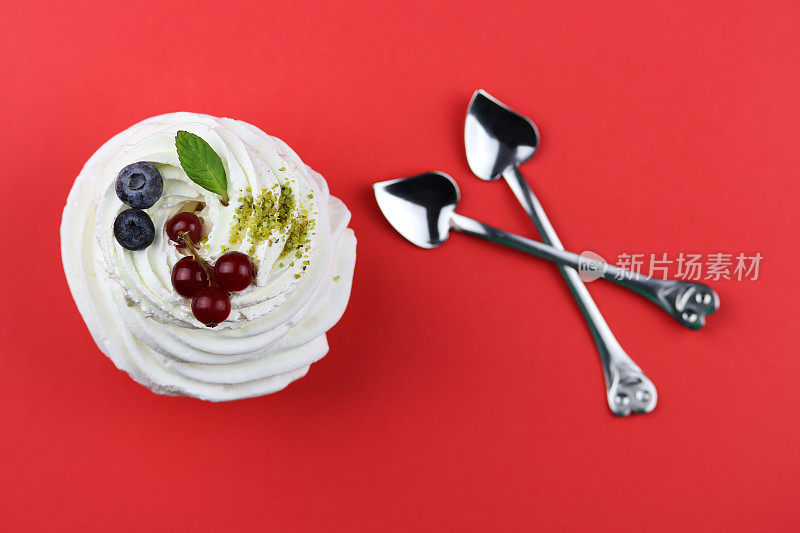 情人节美食。安娜·巴甫洛娃甜点，白色蛋白纸杯蛋糕，新鲜浆果，红色背景的心形勺子。本空间