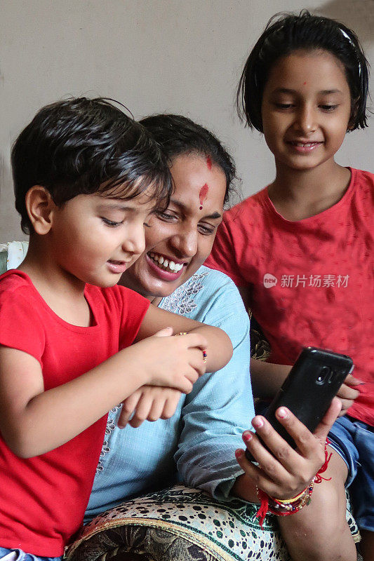 特写图像的印度妇女视频在智能手机上打电话，年轻的女孩在大腿上，大女儿站在后面，妇女穿着Kameez(束腰外衣)传统服装在家庭房间，头部，重点在前景