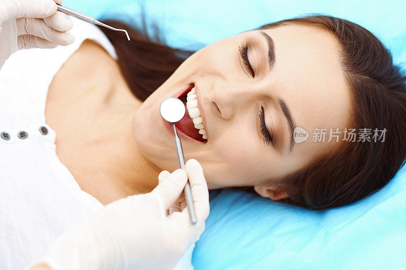 年轻女性病人前往牙科诊所。美丽的女人与健康的直白牙齿坐在牙科椅张开嘴在口腔检查期间，而医生在牙齿工作