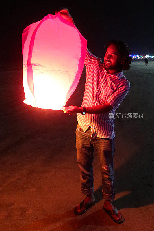 印度男子手持点燃的中国纸灯笼，在海边放纸灯笼的特写，聚焦于前景