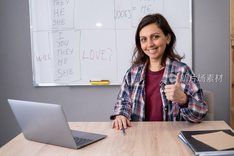 女老师和学生使用笔记本电脑进行在线会议。女教师微笑着翘起大拇指，坐在课桌前，对课堂成绩感到满意