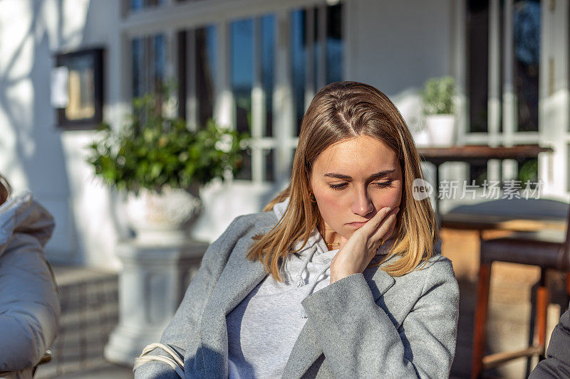 一个金发的年轻女子坐在一家餐馆的露台上晒太阳，脸上带着不高兴的表情。