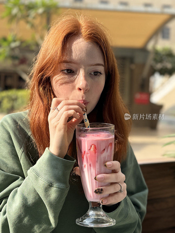 迷人的特写照片，红头发，年轻女子拿着灯罩杯草莓奶昔，用纸吸管喝着不含酒精的饮料，户外咖啡馆餐厅花园，重点在前景