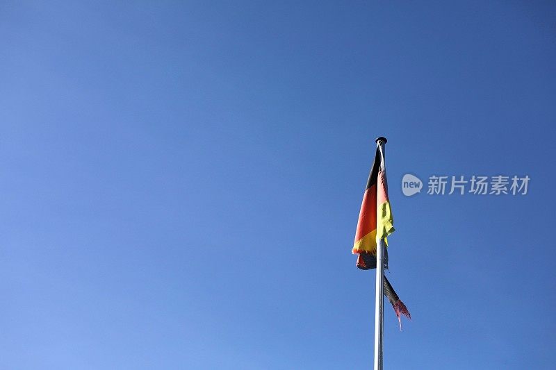 在蔚蓝的天空和开阔的空间前，旗杆上挂着一面破碎的、撕裂的、灾难性的德国国旗