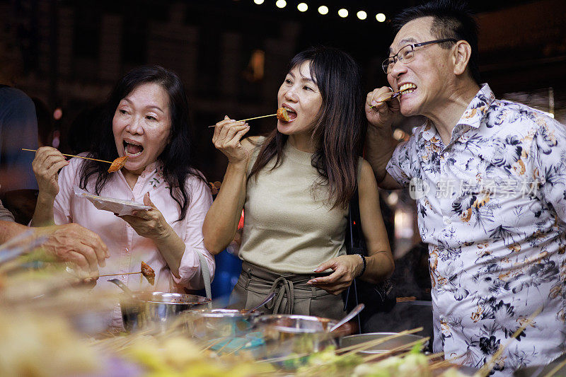 一群亚洲资深游客朋友在槟城乔治城游览时，正在享受经典的街头汽船体验，品尝被称为“乐乐”的各种串。