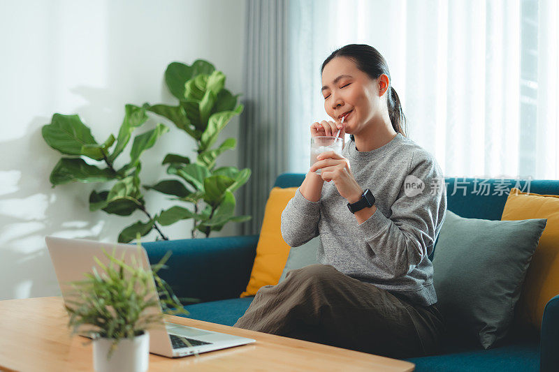 亚洲妇女坐在客厅的沙发上用吸管喝凉水休息。