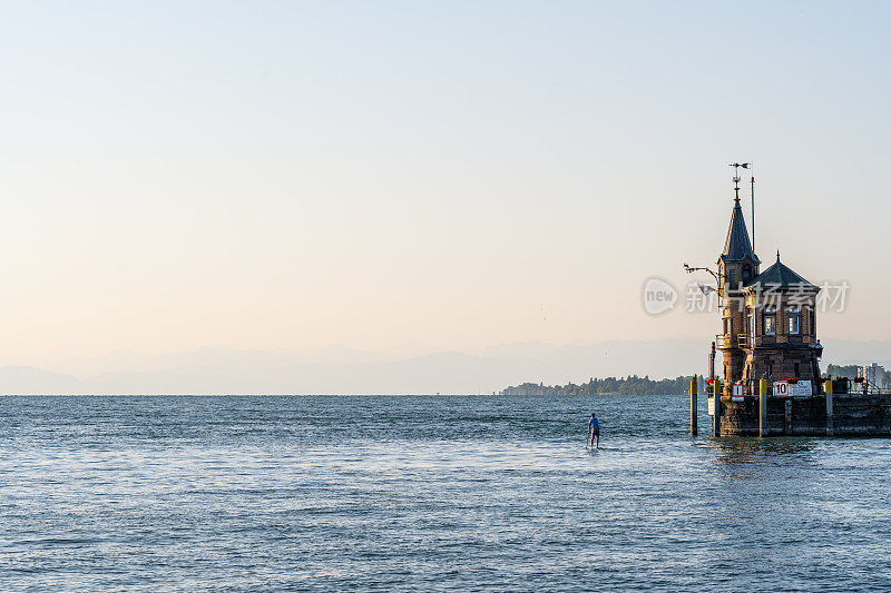 游客站在站立式桨板上经过灯塔和海港入口，入口处挂着康斯坦茨旗。康斯坦茨，康斯坦茨湖，巴登-符腾堡州，德国，欧洲。