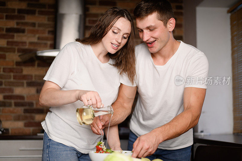 在厨房里，幸福的新婚夫妇准备健康的食物，准备蔬菜沙拉，用油调味，为浪漫的晚餐。男人和女人都试图通过吃素食来过一种健康的生活方式。